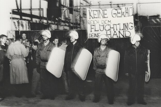Eine Schwaz-weiß-Aufnahme mit vielen Polizisten mit Helmen und Schutzschildern, dahinter halten Demonstranten und Demonstrantinnen ein Transparent