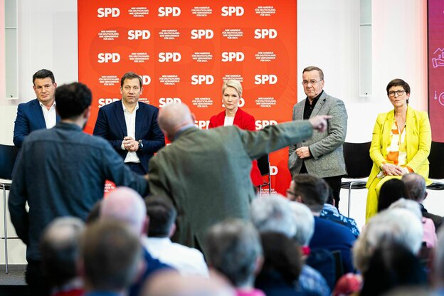 Die SPD Führungsriege im Vordergrund scharf, im Vordergrund das Publikum der Parteimitgliederkonferenz für den Koalitionsvertrag, eine von hinten als älter männlich gelesene Person scheint wild nach vorne zu gestikulieren und deutete mit dem Finger nach r