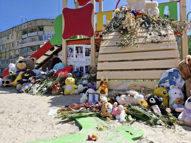 Blumen, Plüschtiere und derlei auf einer Kindertusche auf einem Spielplatz, im Hintergrund typisch sowjetische Plattenbauten in der Ukraine