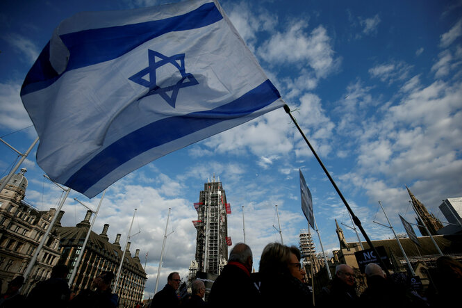 Städtische Szene vor leicht bewölktem Himmel, Menschen auf einer Demonstration, die Flagge Israels wird geschwenkt
