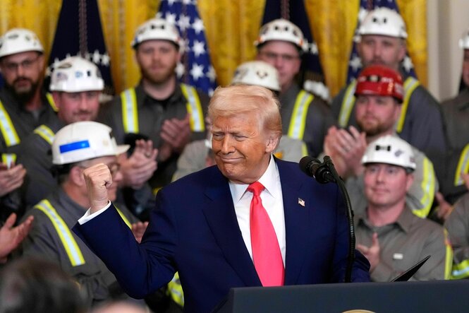 Donald Trump lächelt zurfrieden und ballt die Faust seiner linken Hand, im Hintergrund Männer mit Schutzhelmen auf dem Kopf