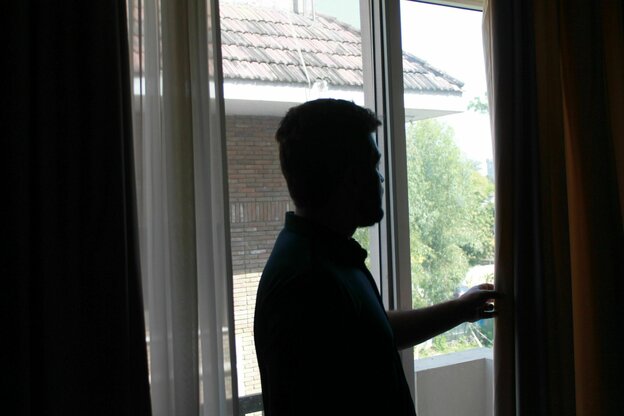 Ein Mann steht am Fenster das teilwiese von einem Vorhang verdeckt wird.