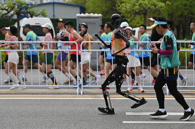 Ein menschlich anmutender Roboter läuft auf der einen Seite eines Absperrgitters, ein Pulk Menschen auf der anderen Seite