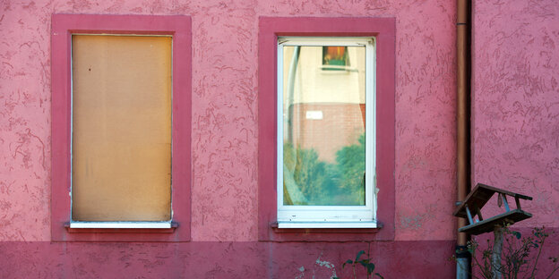 Ein rosafarbenes Haus mit zwei Fenstern, von denen eins mit einer Holzplatte geschützt ist.