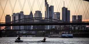 Hinter einer filigranen Brücke erhebt sich die Skyline von Frankfurt a.M., zwei Ruderer im Vordergrund