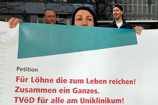 Angestellte des Charité Facility Managements (CFM) stehen bei einer Demonstration für die Eingliederung in den Tarifvertrag des öffentlichen Dienstes (TVöD) hinter einem Plakat