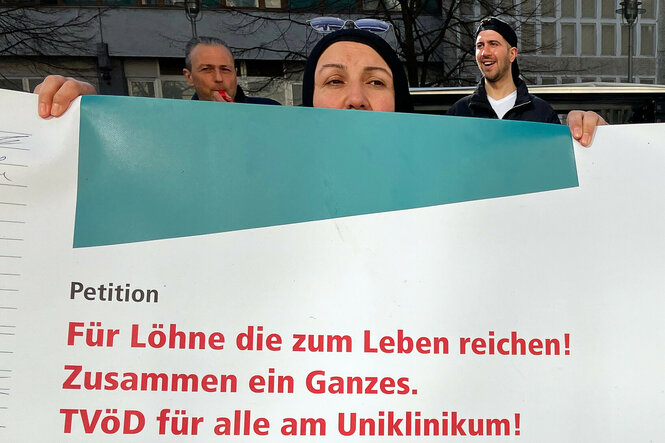 Angestellte des Charité Facility Managements (CFM) stehen bei einer Demonstration für die Eingliederung in den Tarifvertrag des öffentlichen Dienstes (TVöD) hinter einem Plakat