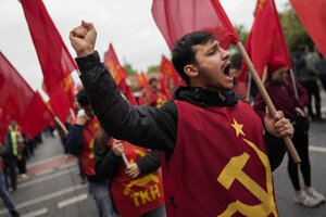 ein Mann reckt die Faust und ruft mit erhobener Fahne etwas, er trägt einen roten Überwurf mit Hammer und Sichel