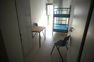 Ein Stockbett, ein Tisch, Schrank und Stuhl aufLinoleumboden in einem zellenähnlichen Raum