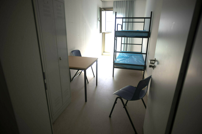 Ein Stockbett, ein Tisch, Schrank und Stuhl aufLinoleumboden in einem zellenähnlichen Raum