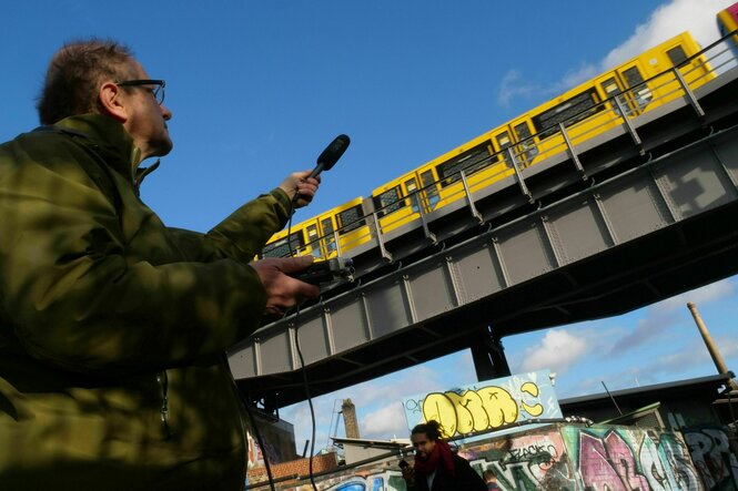 Eine U-Bahn fährt auf einer Brücke, ein Mann im grünen Parka hält ein Mikrofon hoch: Jazzmusiker Paul Brody im Gleisdreieckpark bei der Arbeit