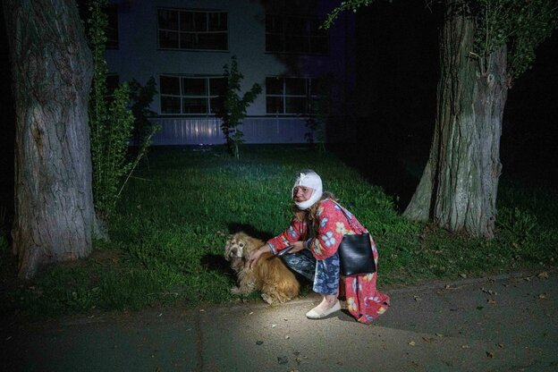 Eine Frau mit Kopfverband kniet neben einem Hund, sie hält ein Handy in der Hand, es ist dunkel