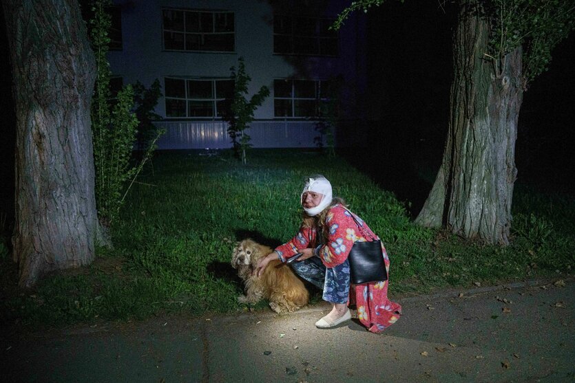Eine Frau mit Kopfverband kniet neben einem Hund, sie hält ein Handy in der Hand, es ist dunkel