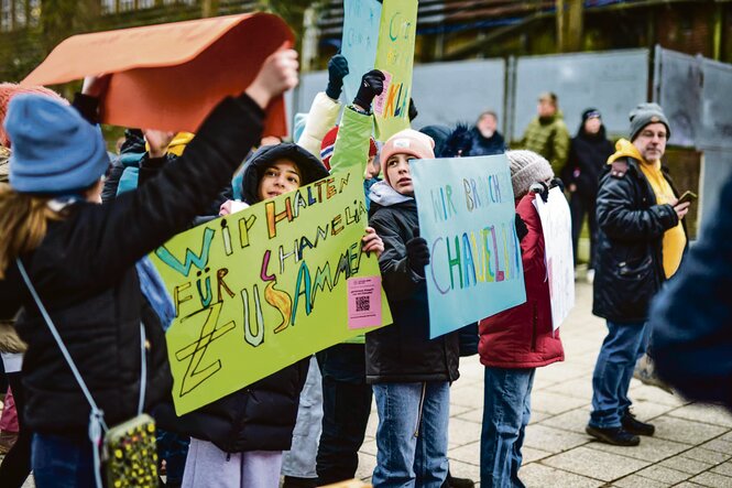 Mehrere Kinder stehen bei einer Demonstration im Freien und halten bunte Plakate mit handgeschriebenen Botschaften wie „Wir halten für Chanelia zusammen“ und „Wir brauchen Chanelia“ hoch.
