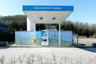 Eine Wasserstofftankstelle mit einer Zapfsäule ohne Auto oder LKW und einem bunten Plakat auf dem gemalte Windkrafträder zu sehen sind