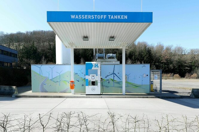 Eine Wasserstofftankstelle mit einer Zapfsäule ohne Auto oder LKW und einem bunten Plakat auf dem gemalte Windkrafträder zu sehen sind