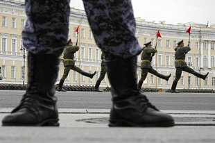 Vier russische Soldaten marschieren mit langgestreckten Beinen und roten Flaggen auf ihrem Bajonett - im Vordergrund sieht man die Steifel und Beine eines Soldaten
