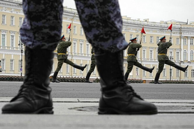 Vier russische Soldaten marschieren mit langgestreckten Beinen und roten Flaggen auf ihrem Bajonett - im Vordergrund sieht man die Steifel und Beine eines Soldaten