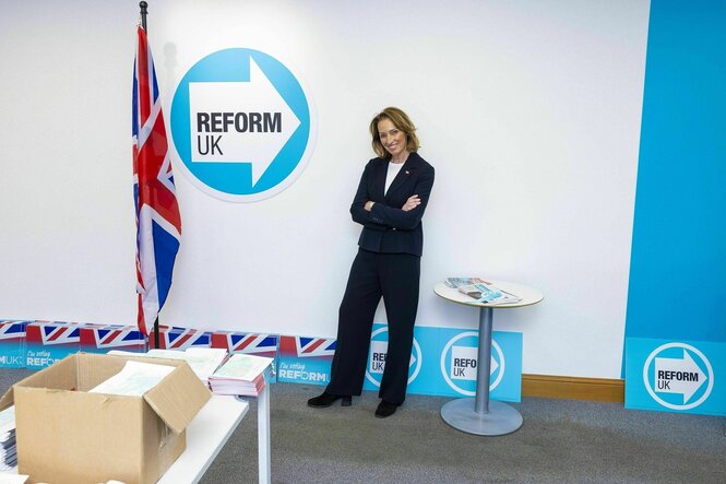 Sarah Pochin steht vor einem Logo in helblau " Reform UK" , der britischen Flagge und ein Kiste mit Unterlagen die auf einem Tisch steht
