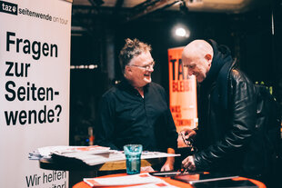 Zwei mittelalte Männer im Gespräch an einem Infostand der „taz seitenwende on tour“. Zwischen Flyern, Roll-up und Plakat entsteht eine offene, einladende Atmosphäre.