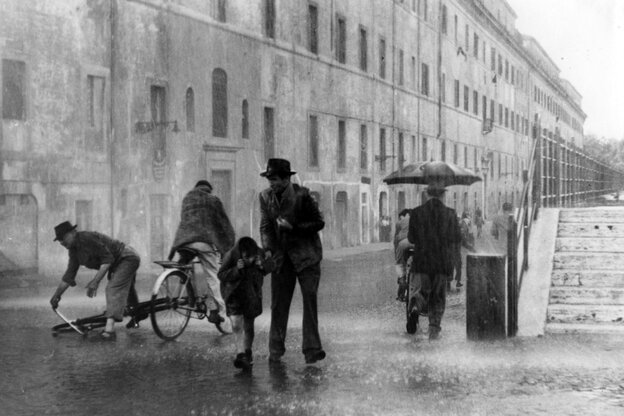 Straßenszene aus einem Schwarz-Weiß-Film: Es regnet in Strömen. Ein Mann und ein Kind laufen durch den Regen und halten sich die Jacken über den Kopf. Hinter ihnen hebt ein Mann ein umgegipptes Fahhred auf, ein weiterer Mann fährt auf einem Fahhrad durch
