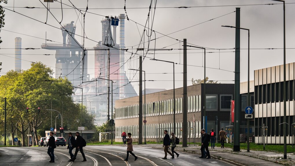Menschen laufen im Gegenlicht über STraßenschienen, im Hintergrund raucht ein Stahlwerk