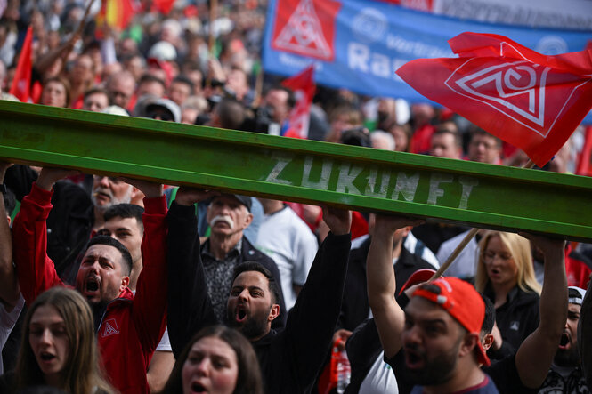 Protestierende Arbeiter unter einer grün gestrichenen STahlschiene mit der Aufschrift Zukunft, IGM Metall Fahnen wehen