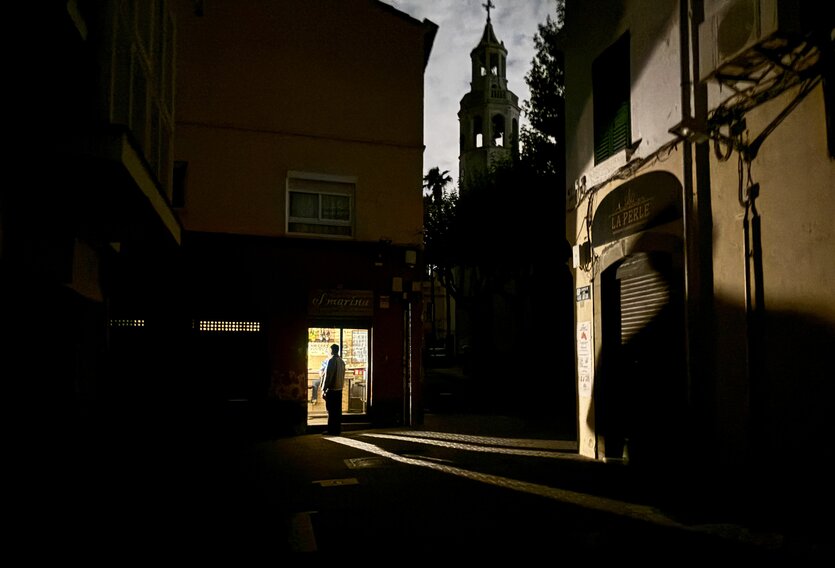 Dunkelheit in einer Gasse, ein Mensch steht vor einem erleuchteten Geschäft, eine Kirche im Hintergrund