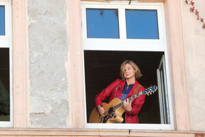 Aus einem geöffneten Fenster in eine höheren Stockwerk schaut die Sängerin Bernadette La Hengst, die Gitarre im Anschlag, auf die Betrachtenden und singt