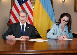 Ein Mann und eine Frau sitzen an einem Schreibtisch und unterzeichnen ein Dokument. Im Hintergrund sind die Flaggen der USA und der Ukraine zu sehen.