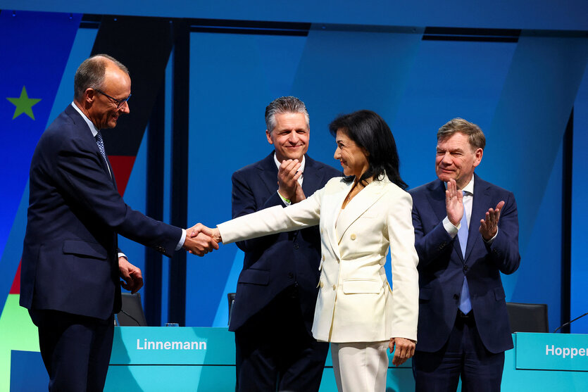 Friedrich Merz und Katherina Reiche stehen auf einer Bühne und schütteln die Hände, im Hintergrund applaudieren zwei Männer