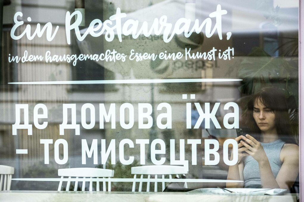 Eine junge Frau sitzt mit ihrem Handy in der Hand in einem Restaurant hinter der Schaufensterscheibe