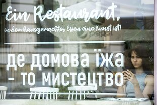 Eine junge Frau sitzt mit ihrem Handy in der Hand in einem Restaurant hinter der Schaufensterscheibe