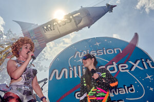 Zwei bunt gekleidete Menschen stehen auf einer Bühne, dahiner eine Rakete mit der Aufschrift 