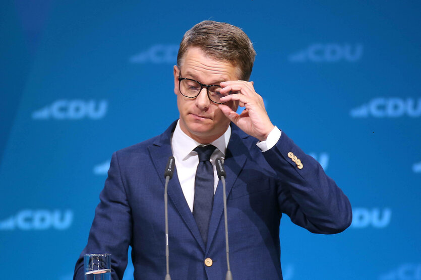 Ein Mann steht an einem Rednerpult und hält eine Hand an seine Brille