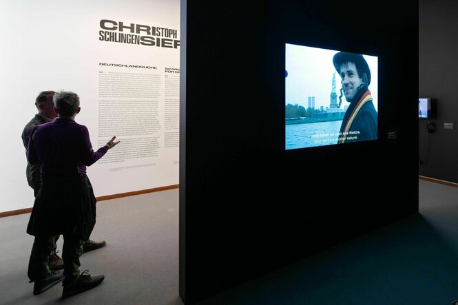 Besucher der Neuen Nationalgalerie stehen in der Ausstellung «Deutschland '99» von Christoph Schlingensief, rechts läuft in Endlosschleife das Video zur Dokumentation der Aktion «Deutschland versenken» aus dem Jahr 1999