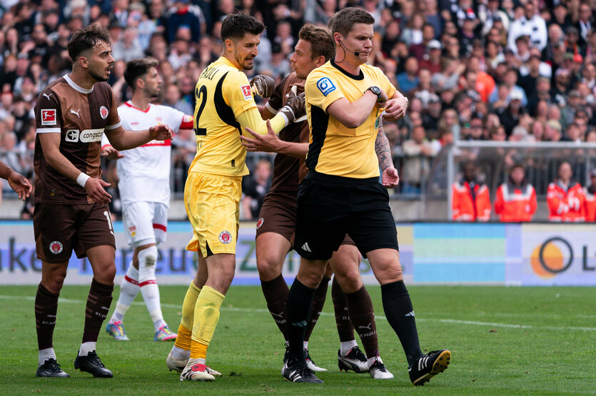 ein Mitspieler stellt sich zwischen St. Paulis Torwart Nikola Vasilj und Schiedsrichter Florian Exner
