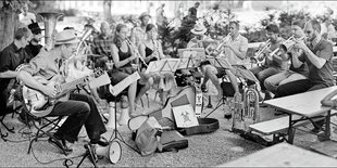 junge leute mit gitarre, banjo und vielen blasinstrumenten sitzen im biergarten und spielen