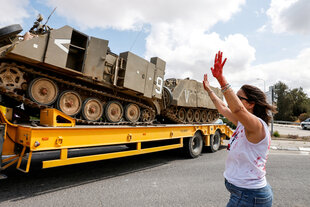 Ein Lastwagen, auf dessen Anhänger Panzer stehen, fährt an einer gestikulierenden Person vorbei