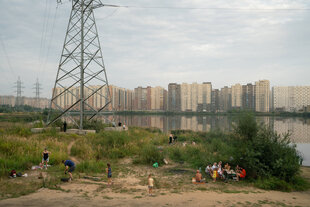 Im Hintergrund eine moderne Wohnsiedlung, davor ein See. Unter einem Strommasten machen Leute am Ufer Picknick