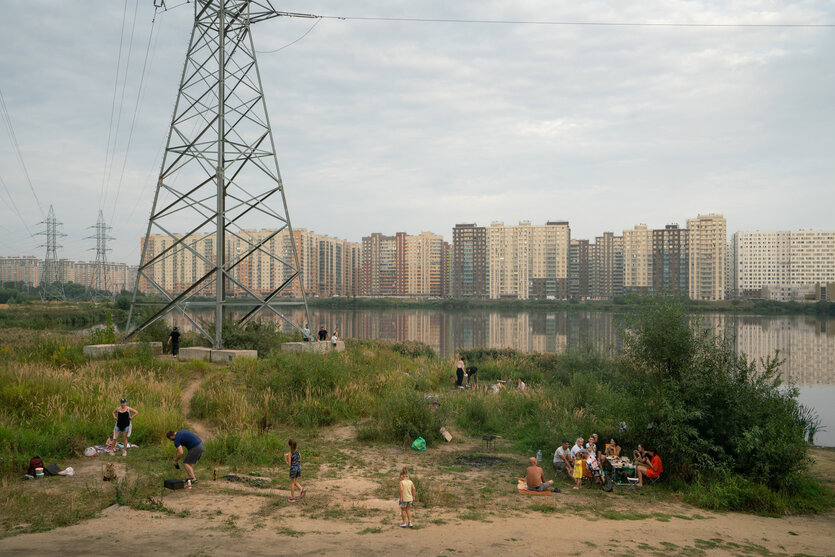 Im Hintergrund eine moderne Wohnsiedlung, davor ein See. Unter einem Strommasten machen Leute am Ufer Picknick