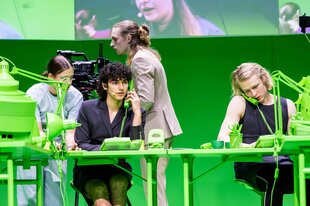 Junge Berliner Schauspieler:innen sitzen am Nachrichtendesk in einer Szene von Newsroom. Möbel und Wände sind grün