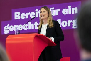 Clara Bünger steht vor einer pinken Wand und hinter einem roten Pult und hält eine Rede.