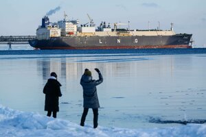 Ein Tanker mit LNG auf der Seite gepinselt im Fokus, im Vordergrund zwei Personen in dicken Jacken und auf Eis stehend