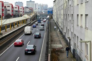 Eine zweispurige Autobahn führt rechts an einer grauen Häuserzeile vorbei, links die Haltestelle der Straßenbahn, die Gegenfahrbahn und Wohnhäuser im Stil der 70er Jahre