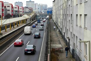 Eine zweispurige Autobahn führt rechts an einer grauen Häuserzeile vorbei, links die Haltestelle der Straßenbahn, die Gegenfahrbahn und Wohnhäuser im Stil der 70er Jahre