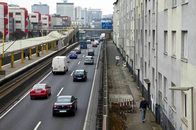 Eine zweispurige Autobahn führt rechts an einer grauen Häuserzeile vorbei, links die Haltestelle der Straßenbahn, die Gegenfahrbahn und Wohnhäuser im Stil der 70er Jahre
