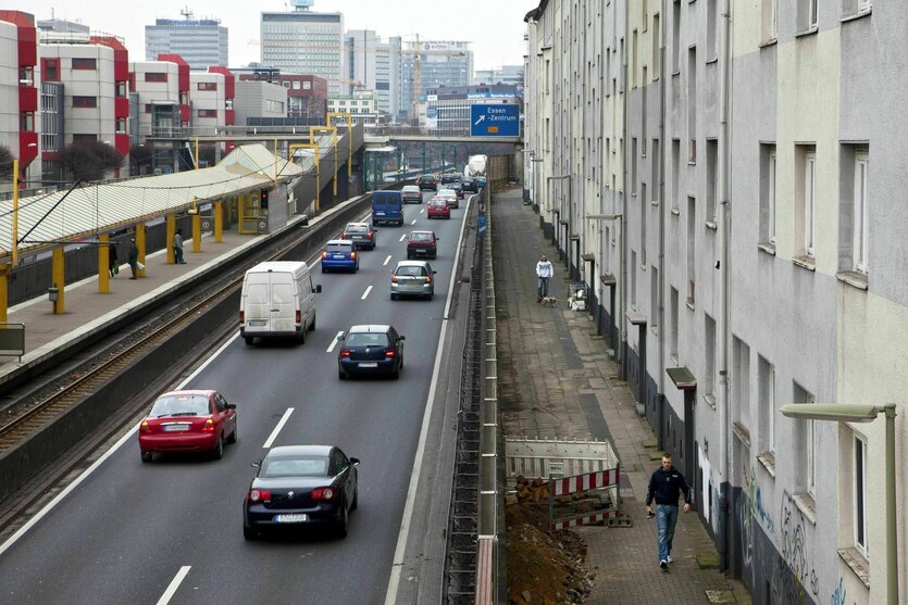 Eine zweispurige Autobahn führt rechts an einer grauen Häuserzeile vorbei, links die Haltestelle der Straßenbahn, die Gegenfahrbahn und Wohnhäuser im Stil der 70er Jahre