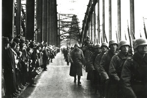Soldaten der Wehrmacht in Uniform mit Helm marschieren über die Rheinbrücke. Dabei werden sie von vielen Zuschauern auf der anderen Seite der Brücke beobachtet