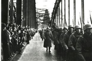 Soldaten der Wehrmacht in Uniform mit Helm marschieren über die Rheinbrücke. Dabei werden sie von vielen Zuschauern auf der anderen Seite der Brücke beobachtet